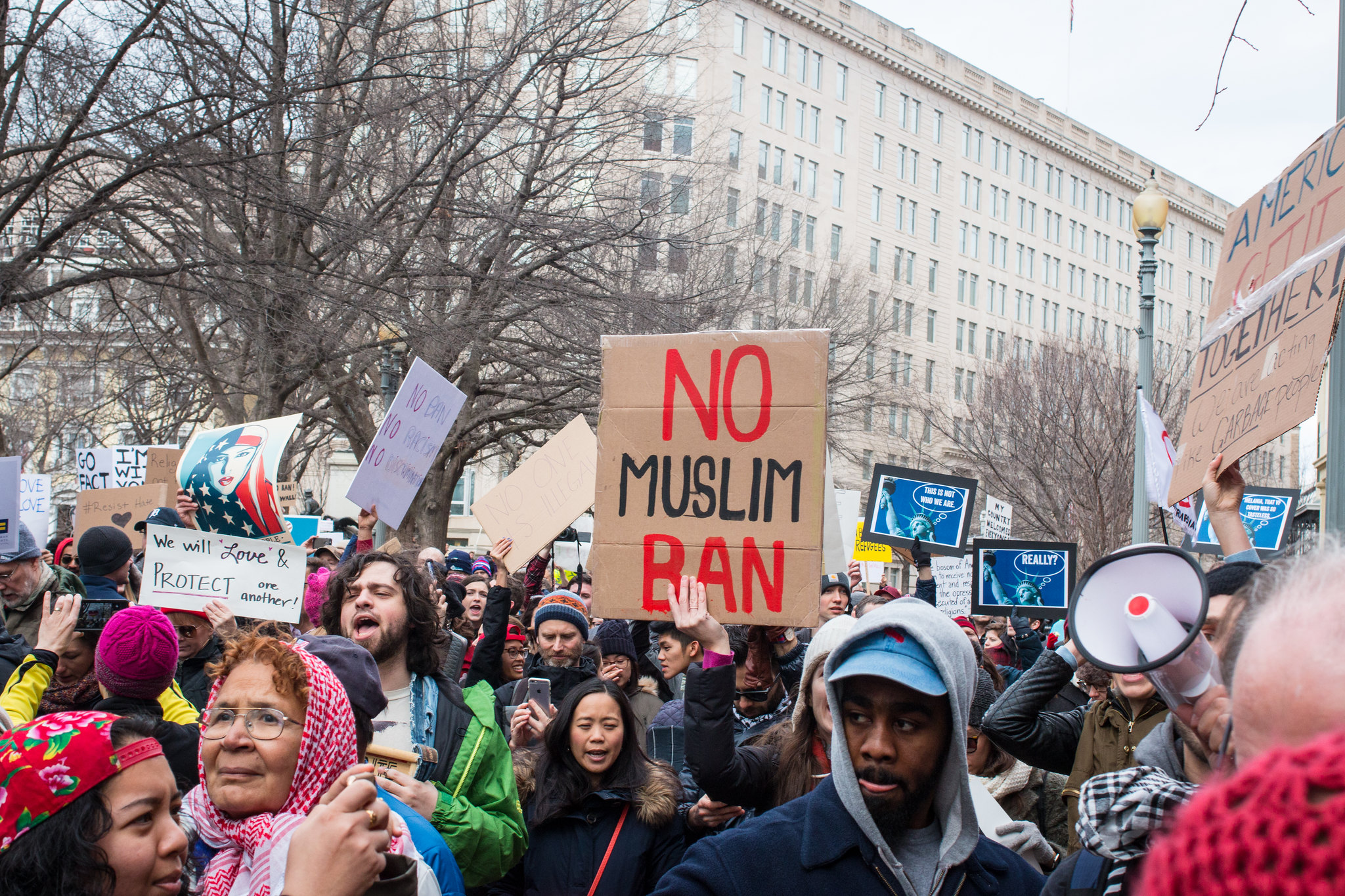 Multiracial crowd in front of what looks like a government building; a prominent hand-made sign says "No Muslim Ban"