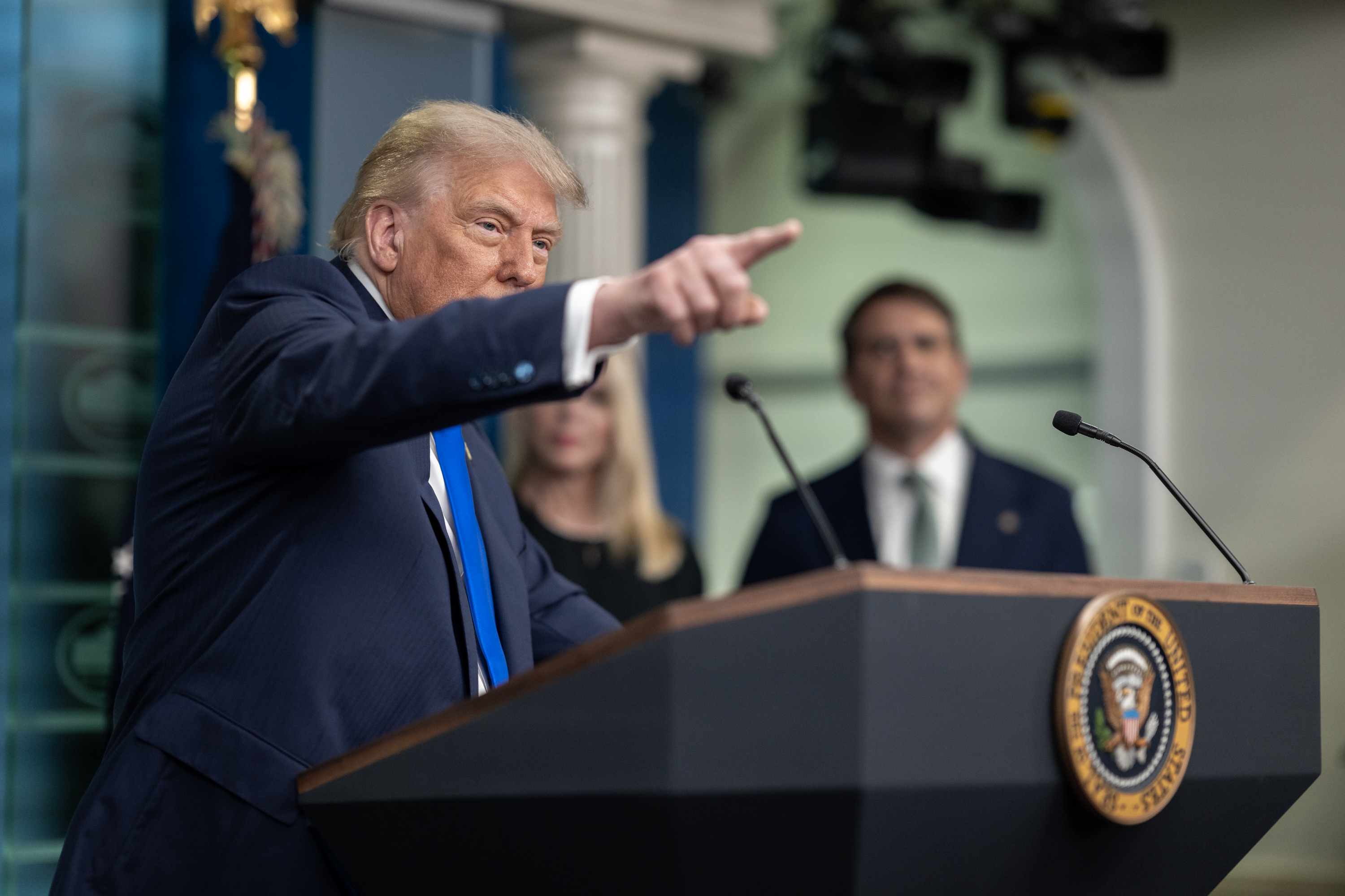 Donald Trump stands behind a podium bearing the seal of the President, pointing with his right arm and hand.