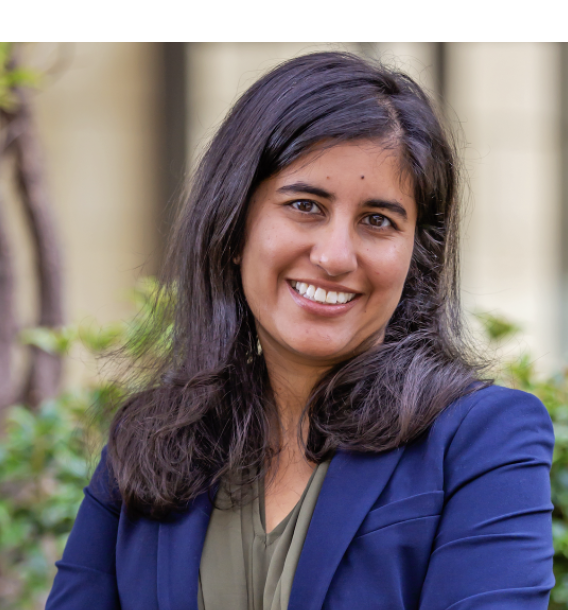 Headshot of Easha Anand, in a blue blazer and green shirt, with crossed arms, in front of foliage.