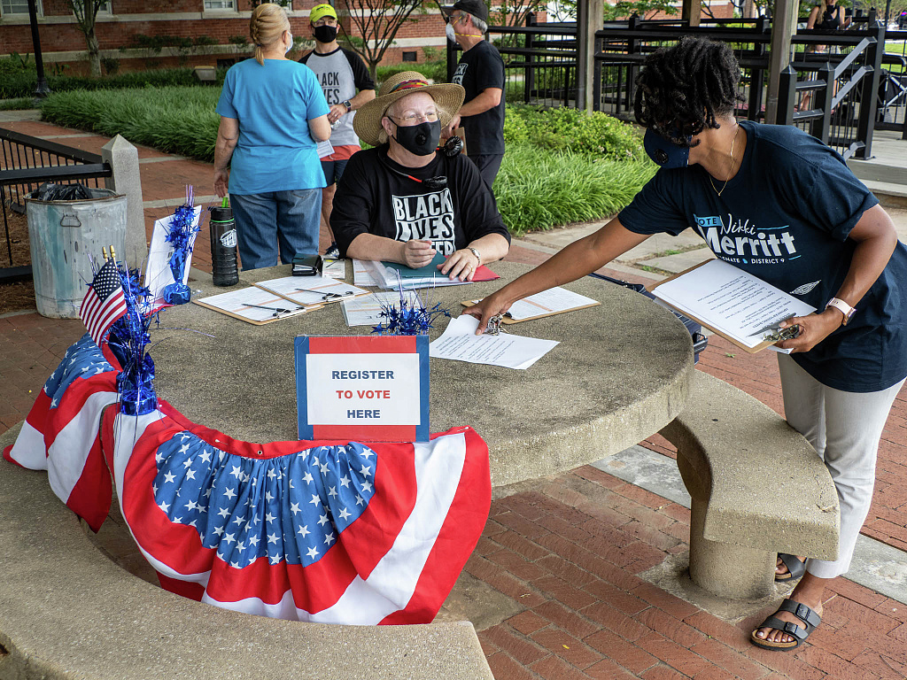 A woman wearing a mask and a Black Lives Matter shirt sits at a table adorned with American flags and a sign that says "Register to Vote Here" while another woman, also in a mask, stands and organizes documents on the table.